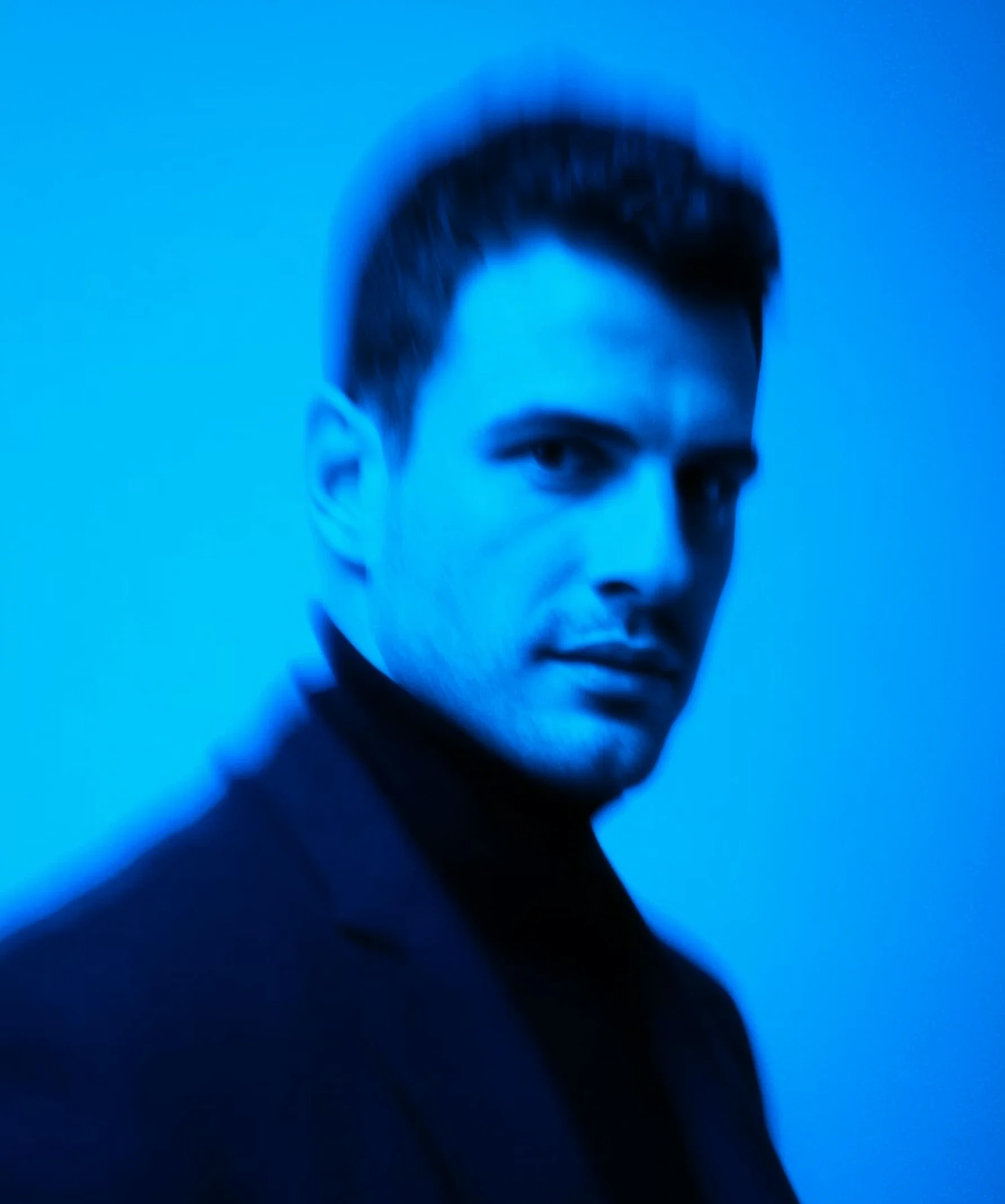 Portrait of a man with short dark hair and a serious expression under blue lighting with a blurred effect.