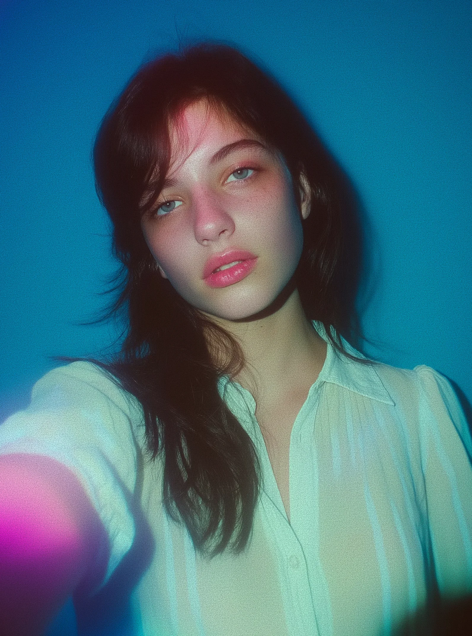 Young woman with dark hair and blue eyes wearing a light-colored buttoned blouse against a blue background.